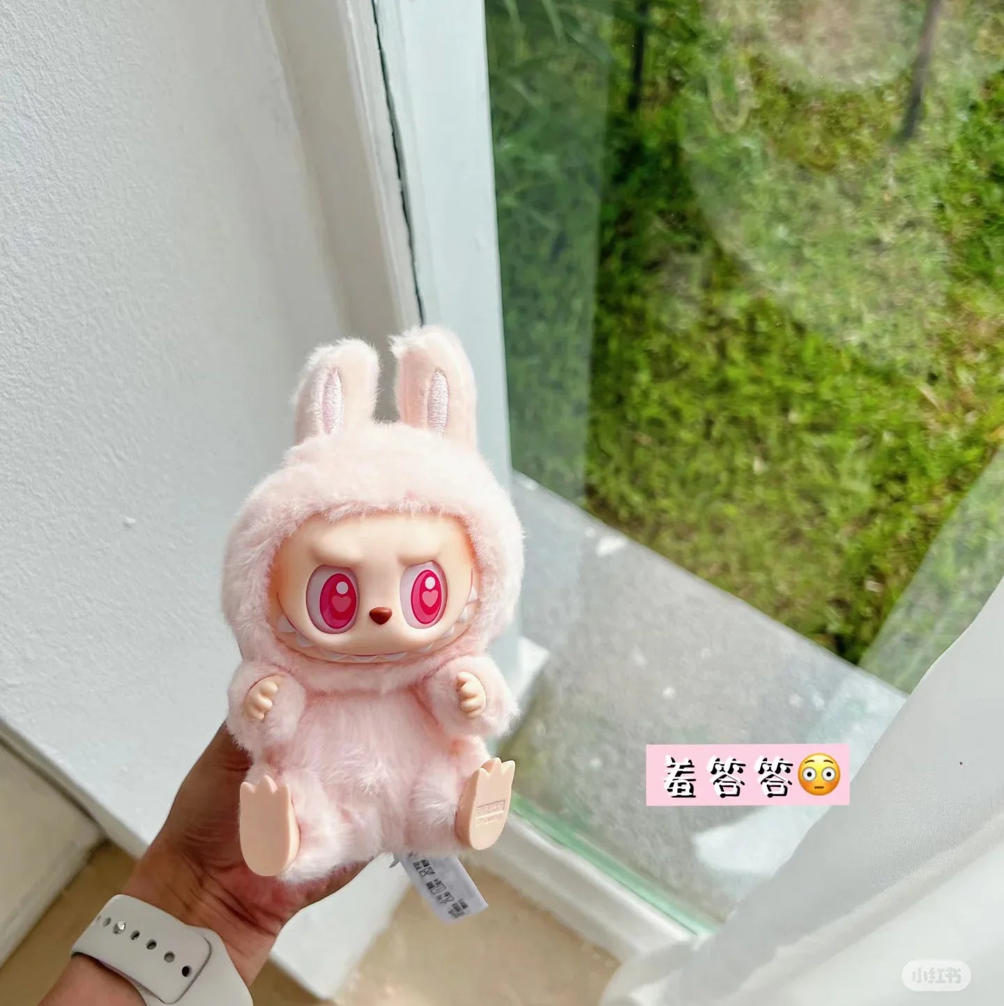 Plush toy Labubbu with pink bunny ears held by a hand, with a blurred outdoor background.