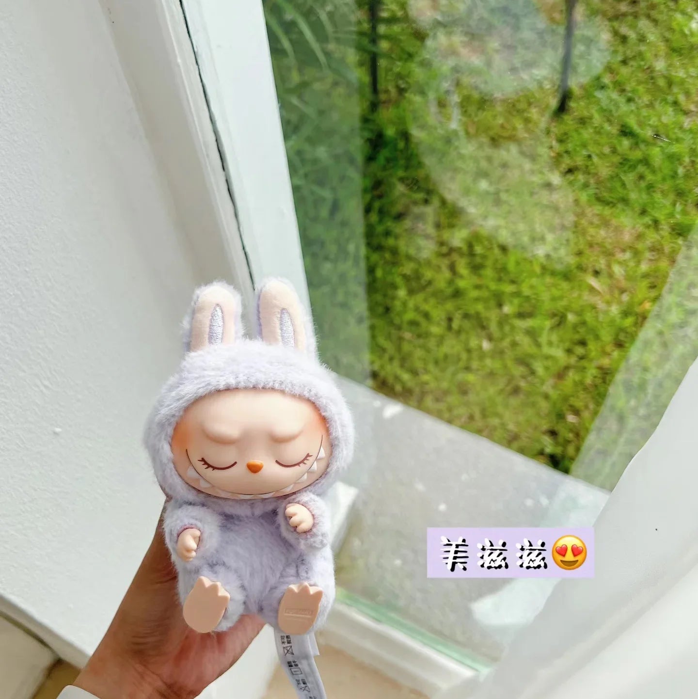 Plush toy Labubu in a gray outfit held in front of a window with greenery outside.