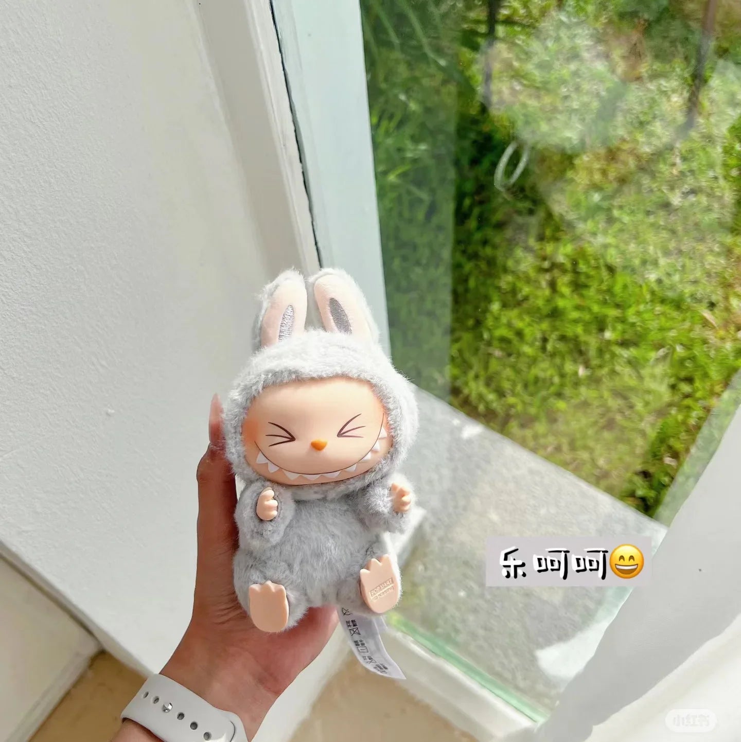 Plush toy with rabbit ears Labubu held in front of a window with greenery outside
