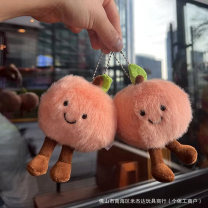 Jellycat. Two plush peach-shaped keychains held by a hand with a blurred background.