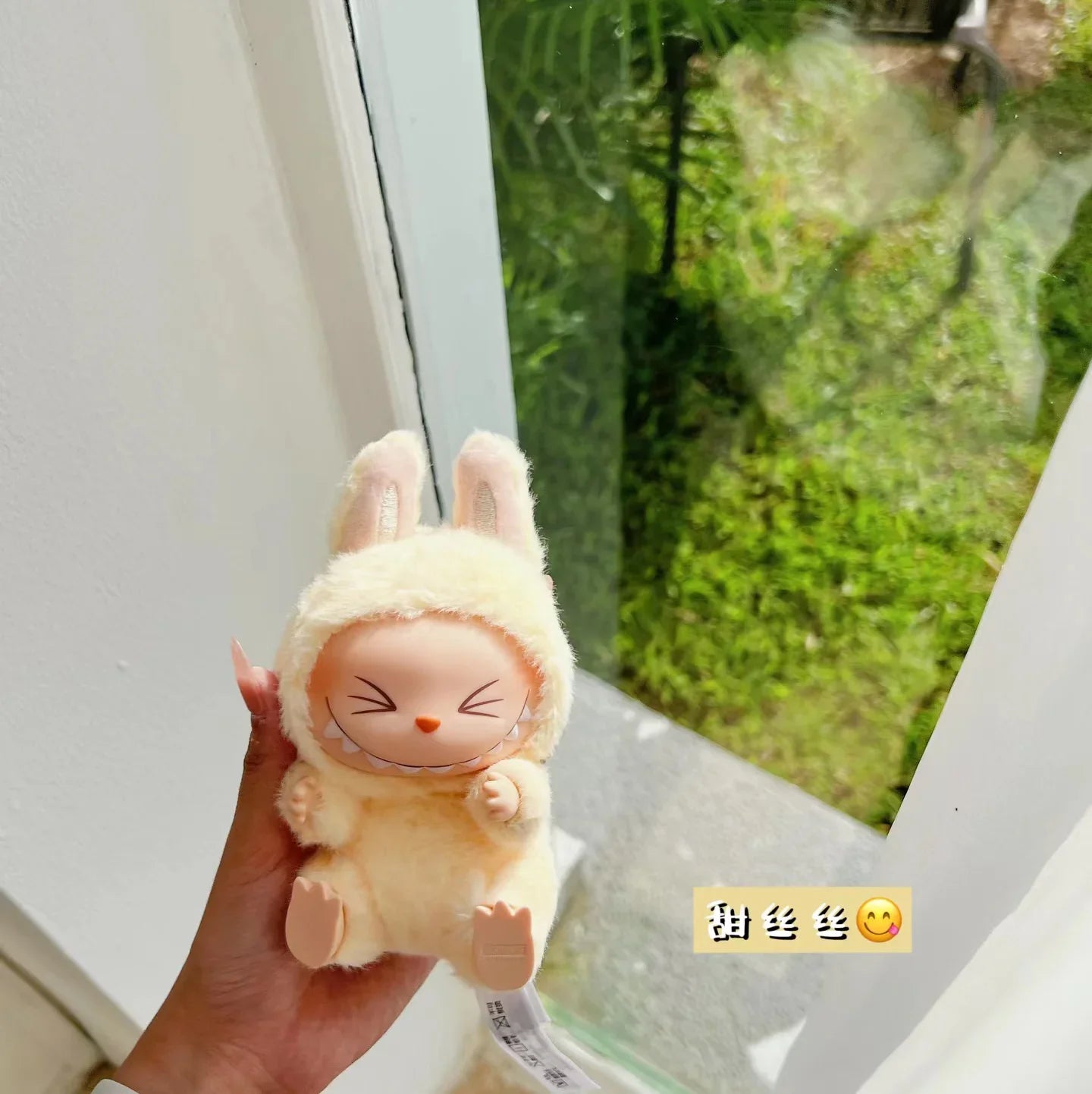 Plush toy resembling a character with bunny ears held by a hand, with a blurred green outdoor background. Labubu have a seat