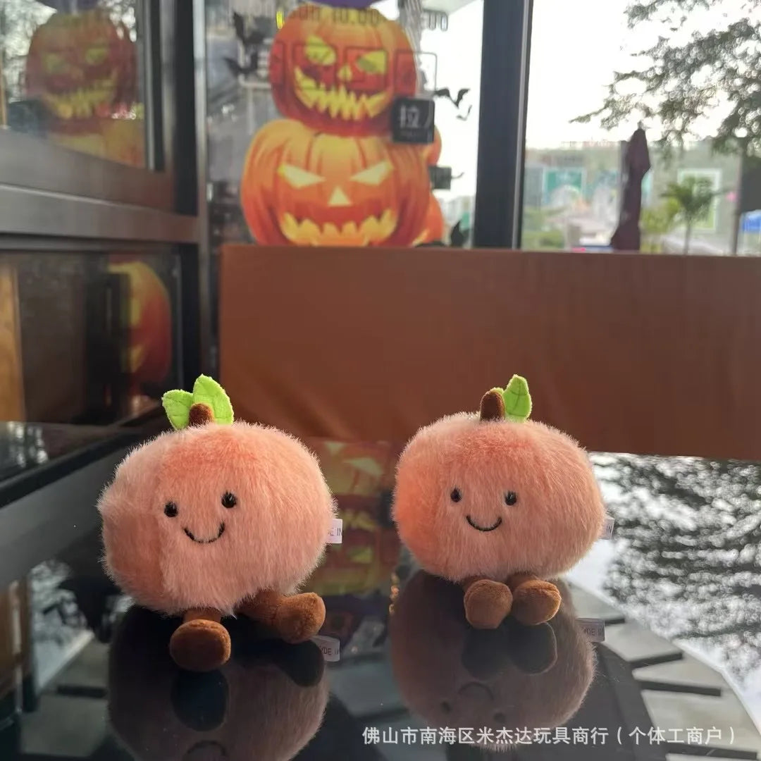 Jellycat. Two plush pumpkins with smiling faces on a reflective surface, with Halloween decorations in the background.